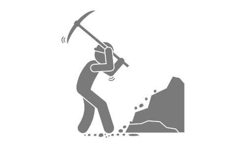 A person wearing safety gear uses a pickaxe to break rocks on the ground, with debris falling around them.