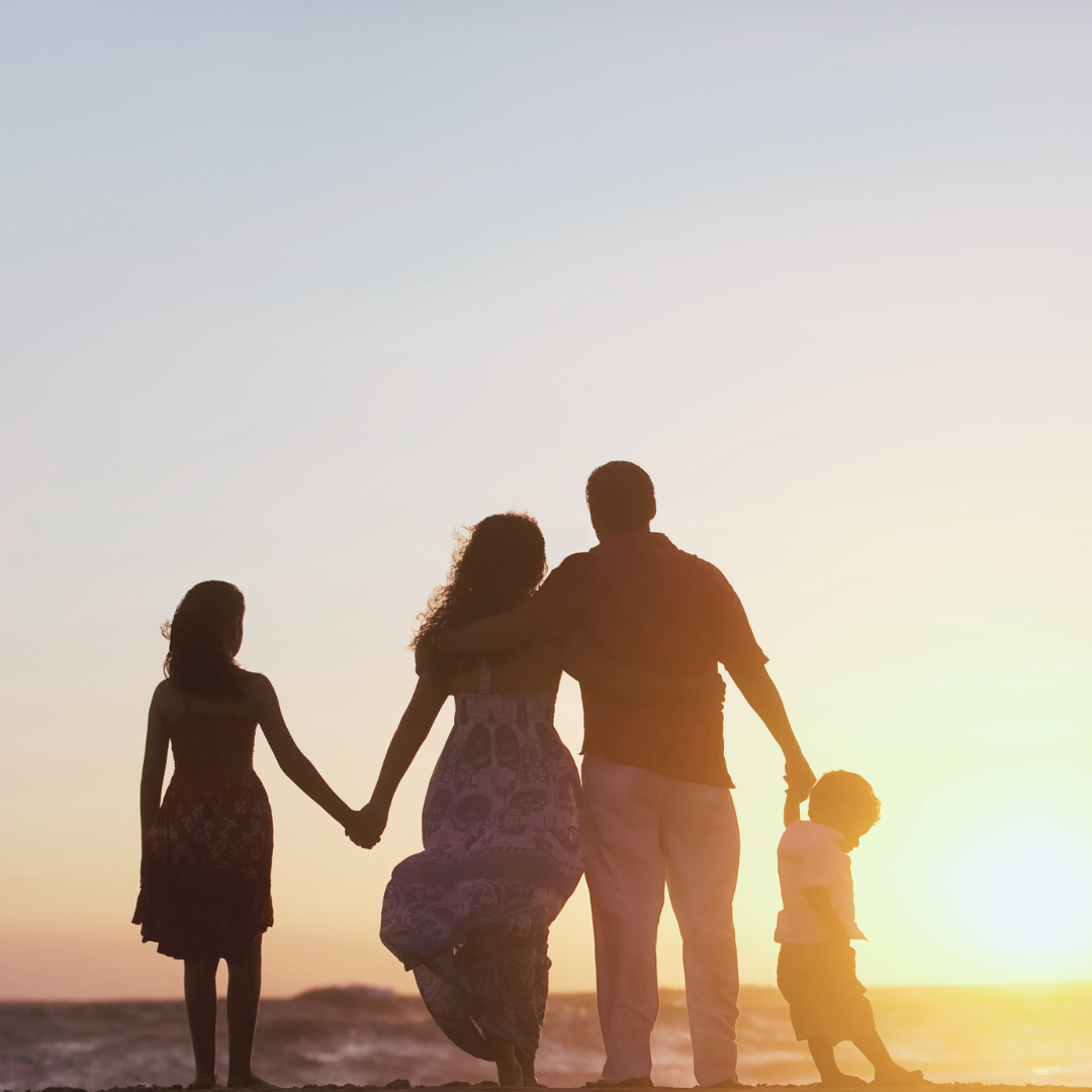 Four people stand hand in hand on a beach, facing the ocean during sunset.